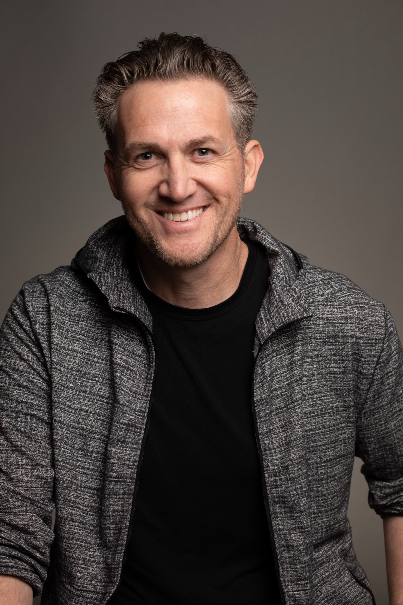 Matthew Dearing, 2025. Grey hoodie, warm smile, grey studio backdrop.